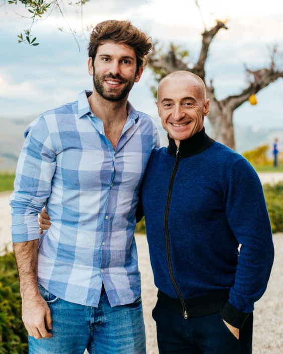 Gianluca Fazio e Juri Chechi sorridono abbracciati all’aperto, in un paesaggio collinare delle Marche al tramonto. Gianluca indossa una camicia a quadri azzurri, Juri una felpa blu con zip. La foto fa parte di una campagna di comunicazione per la Regione Marche, dedicata all’accoglienza, alla bellezza e ai volti del territorio.