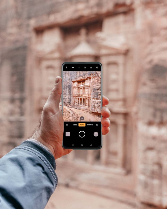 Una mano tiene uno smartphone Oppo mentre fotografa il Tesoro di Petra, in Giordania. Sullo schermo del telefono è perfettamente inquadrata la celebre facciata scolpita nella roccia. L’immagine fa parte di una campagna di comunicazione per Oppo, dedicata alla qualità fotografica del dispositivo in viaggio.
