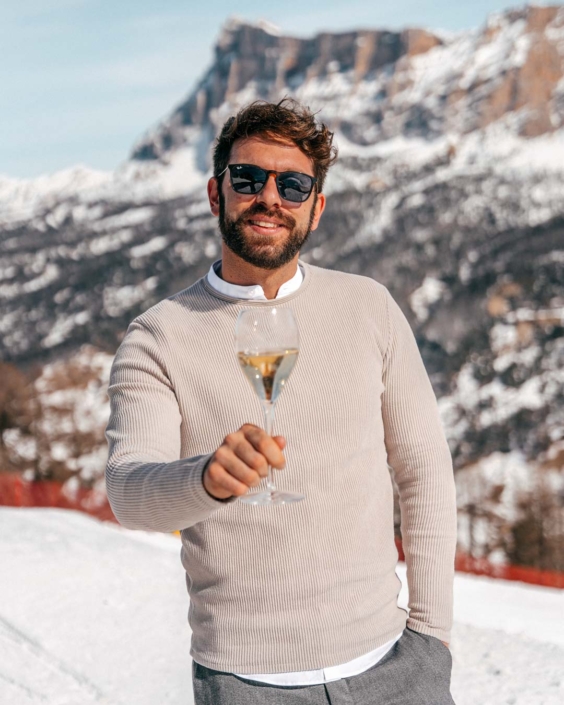 Gianluca Fazio brinda con un calice di Ferrari Trento davanti a un paesaggio alpino innevato. Indossa occhiali da sole e un maglione beige chiaro, con le Dolomiti sullo sfondo. La foto fa parte di una campagna di comunicazione per Ferrari Trento, tra eleganza informale e atmosfera di montagna.