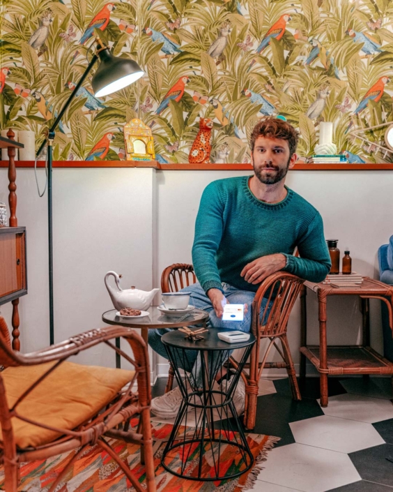 Ritratto ambientato di Gianluca Fazio seduto in un salotto dallo stile eclettico, con carta da parati tropicale e arredi in vimini. Indossa un maglione verde petrolio e jeans, e sta effettuando un pagamento contactless con lo smartphone su un lettore POS. Sulla scena, una teiera bianca, tazze da tè e oggetti decorativi. L'immagine fa parte di una campagna di comunicazione per Mastercard.