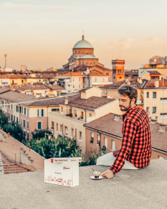 Gianluca Fazio seduto su un tetto al tramonto, mentre sorseggia un caffè Nespresso con vista sui tetti di Bologna. Indossa una camicia a quadri rossi e neri, con accanto una confezione di capsule Nespresso “Piacere”. Sullo sfondo si distingue la cupola della Chiesa del Sacro Cuore. La foto fa parte di una campagna di comunicazione per Nespresso, tra gusto e scorci urbani.