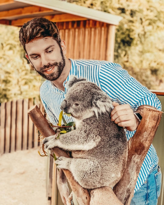 Gianluca Fazio accarezza un koala mentre lo osserva con uno sguardo affettuoso, in un ambiente naturale e curato. Indossa una camicia a righe azzurre e jeans, e si trova accanto a un recinto in legno. L’immagine fa parte di una campagna di comunicazione per Tourism Australia, dedicata alla fauna locale e alle esperienze autentiche nel Paese.