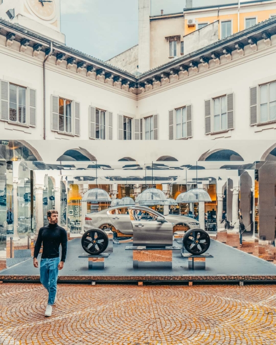 Gianluca Fazio cammina nel cortile di un palazzo storico durante la Milano Design Week 2019, davanti a un’installazione specchiante con la scocca e i componenti di una Jaguar XE. L’opera riflette l’architettura circostante creando un dialogo tra design automobilistico e contesto urbano. La foto fa parte di una campagna di comunicazione per Jaguar.
