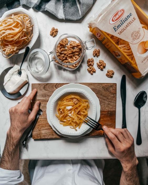 Ripresa dall’alto di un piatto di tagliatelle con ricotta e noci su un tagliere di legno, pronto per essere gustato. Ai lati della scena si vedono le mani di un uomo a tavola, insieme agli ingredienti principali: noci, ricotta fresca e un pacco di Tagliatelle Emiliane Barilla. L’immagine fa parte di una campagna di comunicazione dedicata all’autenticità e alla semplicità della cucina italiana.