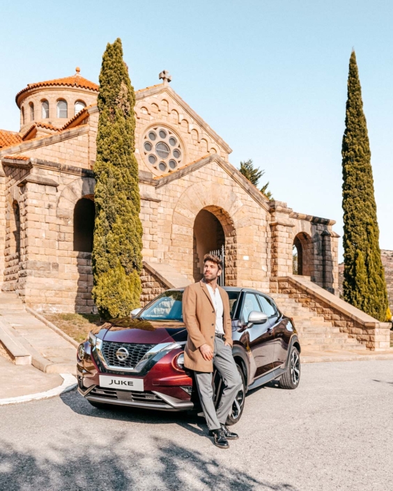 Gianluca Fazio appoggiato a una Nissan Juke parcheggiata davanti a una chiesa romanica in Catalogna. Indossa un cappotto cammello e posa con sguardo rivolto verso l’alto, tra architettura storica e design contemporaneo. L’immagine fa parte di una campagna di comunicazione per Nissan, ambientata tra i paesaggi culturali della Catalogna.