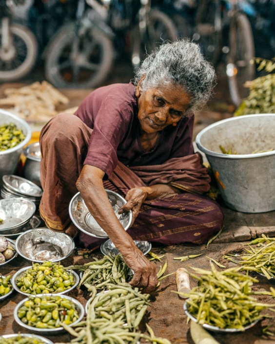 Donna anziana indiana seduta a terra mentre seleziona legumi freschi in un mercato locale. Indossa un sari color prugna e lavora con attenzione circondata da ciotole di metallo colme di verdure. La scena, parte di uno shooting fotografico realizzato in India, cattura un momento di vita quotidiana autentica e intensa.
