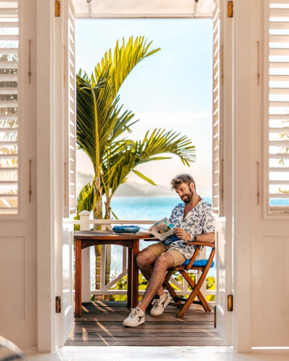 Gianluca Fazio seduto su una veranda affacciata sul mare tropicale, mentre legge una guida di viaggio sulla Giamaica. Indossa una camicia floreale e pantaloncini chiari, circondato da luce naturale e palme. La scena trasmette relax e bellezza esotica, ed è parte di una campagna di comunicazione per l'ente del turismo della Giamaica.
